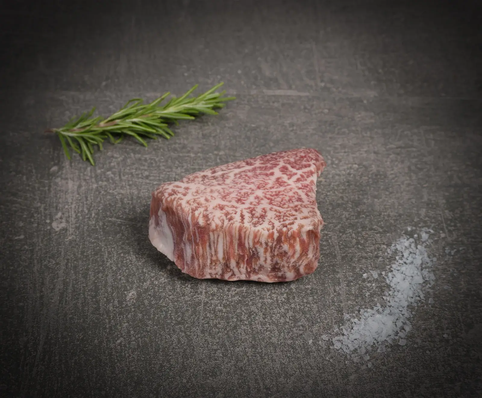 Ein rohes, stark marmoriertes Wagyu-Steak liegt auf einer dunklen Oberfläche, begleitet von einem frischen Rosmarinzweig und einer Prise grobem Meersalz.