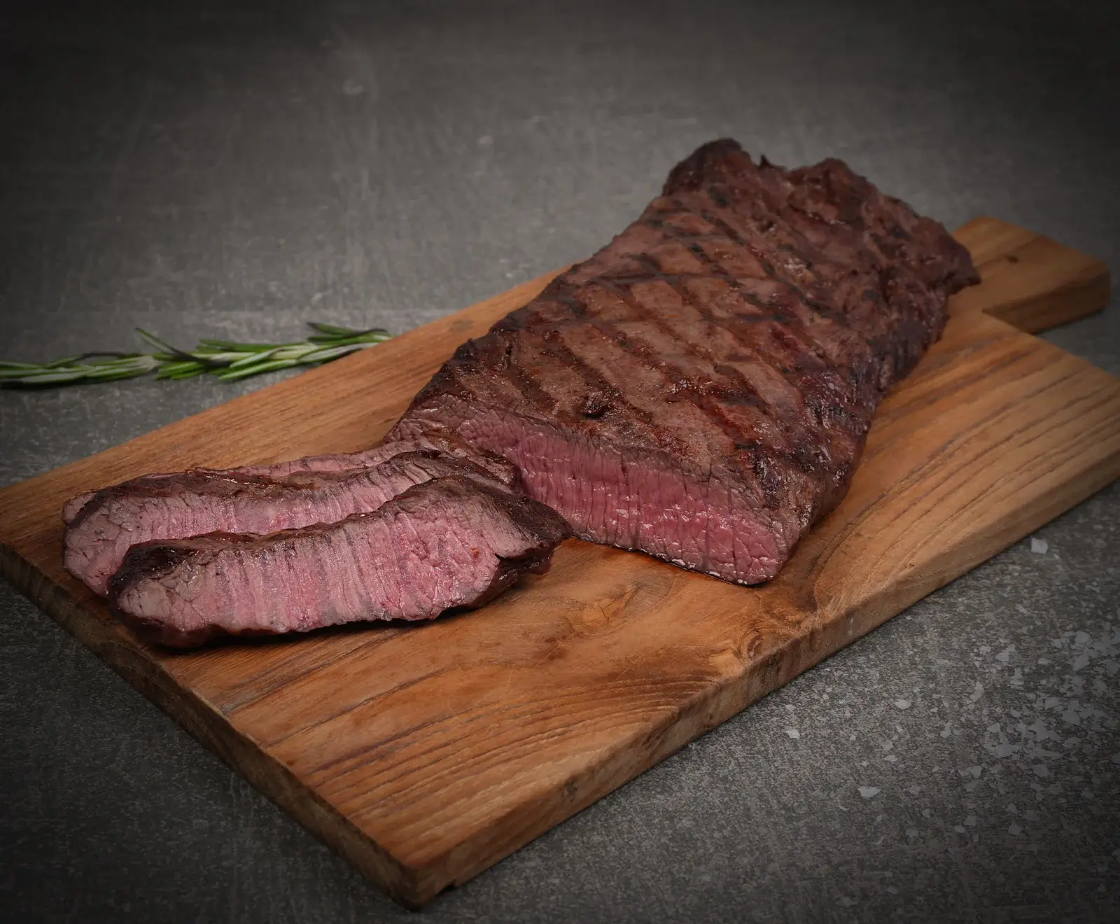Ein gegrilltes Steak, teilweise in Scheiben geschnitten, mit rosa Kern und deutlichen Grillstreifen, liegt auf einem Holzschneidebrett. Rosmarinzweige und Salzkristalle sind auf dunklem Untergrund zu sehen.