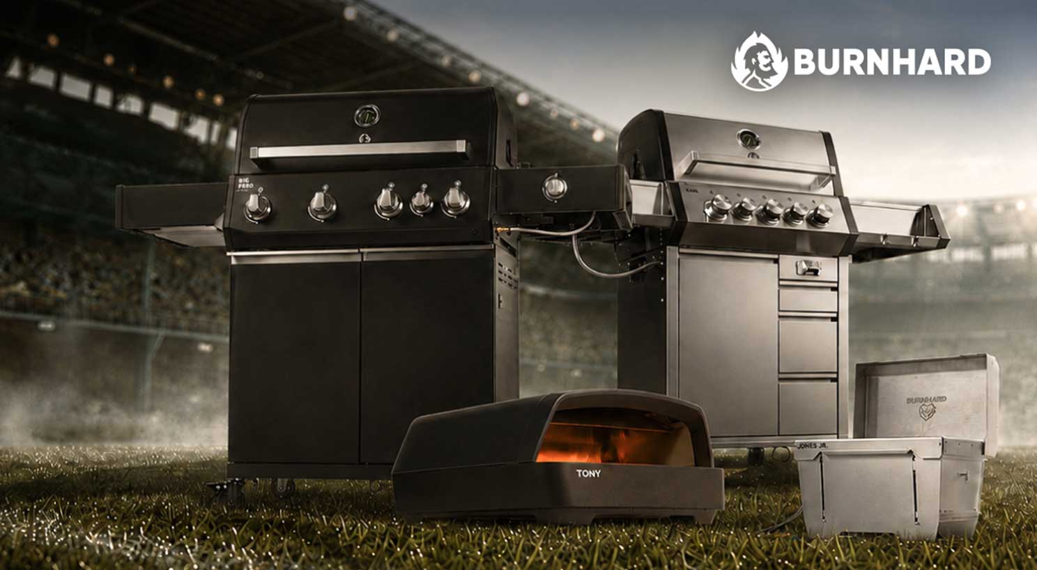 Zwei Burnhard Gasgrills (schwarz "BIG PERO", silber "KARL"), ein Pizzaofen "TONY" und eine Kühlbox "JONES JR." stehen auf Rasen vor einem Stadion. Oben rechts das Burnhard-Logo.