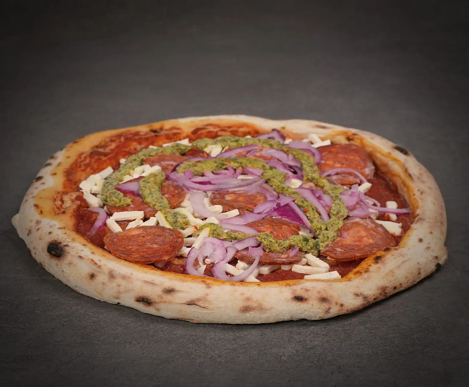 Runde Pizza mit knusprigem Rand, belegt mit Tomatensauce, Salami, weißen Käsestücken, roten Zwiebelringen und grünem Pesto. Die Zutaten sind frisch und der Käse ist noch nicht geschmolzen.