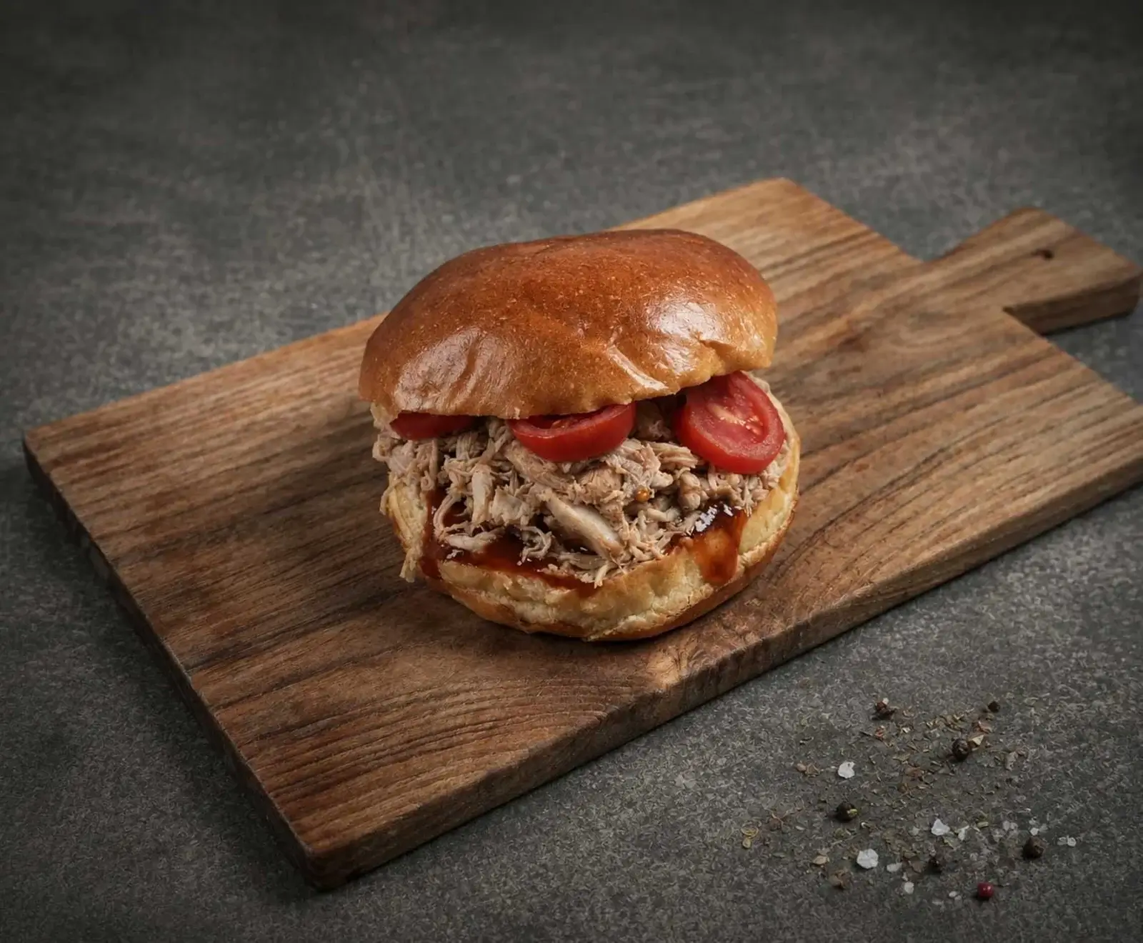 Ein Pulled Pork Burger mit goldbraunem Brötchen, geschreddertem Fleisch, Tomatenscheiben und dunkler Sauce auf einem Holzschneidebrett vor dunklem Hintergrund mit verstreuten Gewürzen.