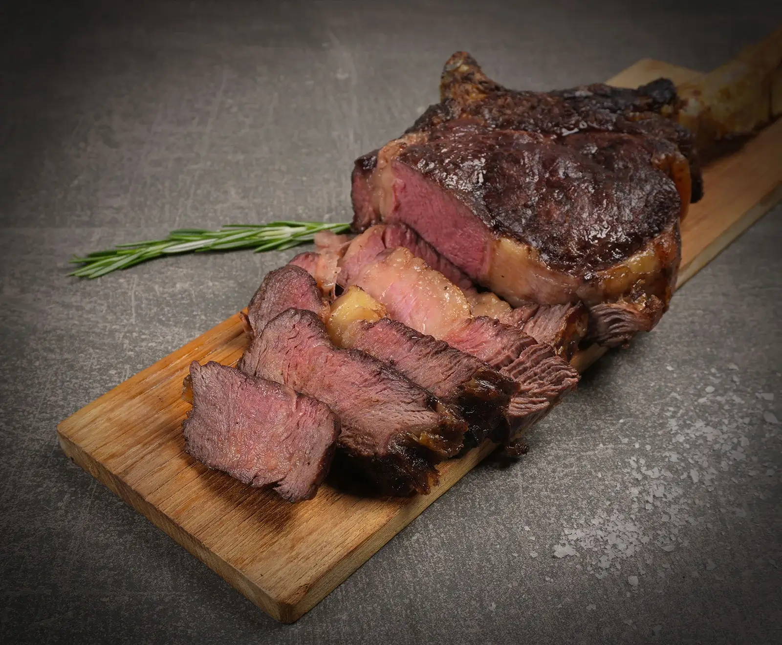 Ein perfekt gegartes, aufgeschnittenes Steak mit knuspriger Kruste und rosa Kern, serviert auf einem Holzbrett. Ein Rosmarinzweig liegt daneben auf dunklem Untergrund mit Salzkristallen.