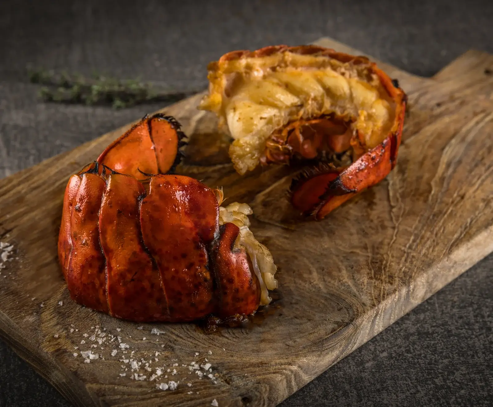 Zwei gegarte Hummerschwänze auf einem rustikalen Holzschneidebrett. Einer ist intakt, der andere ist aufgeschnitten und zeigt das zarte weiße Hummerfleisch. Grobe Salzkristalle liegen auf dem Brett, im Hintergrund ein Kräuterzweig.