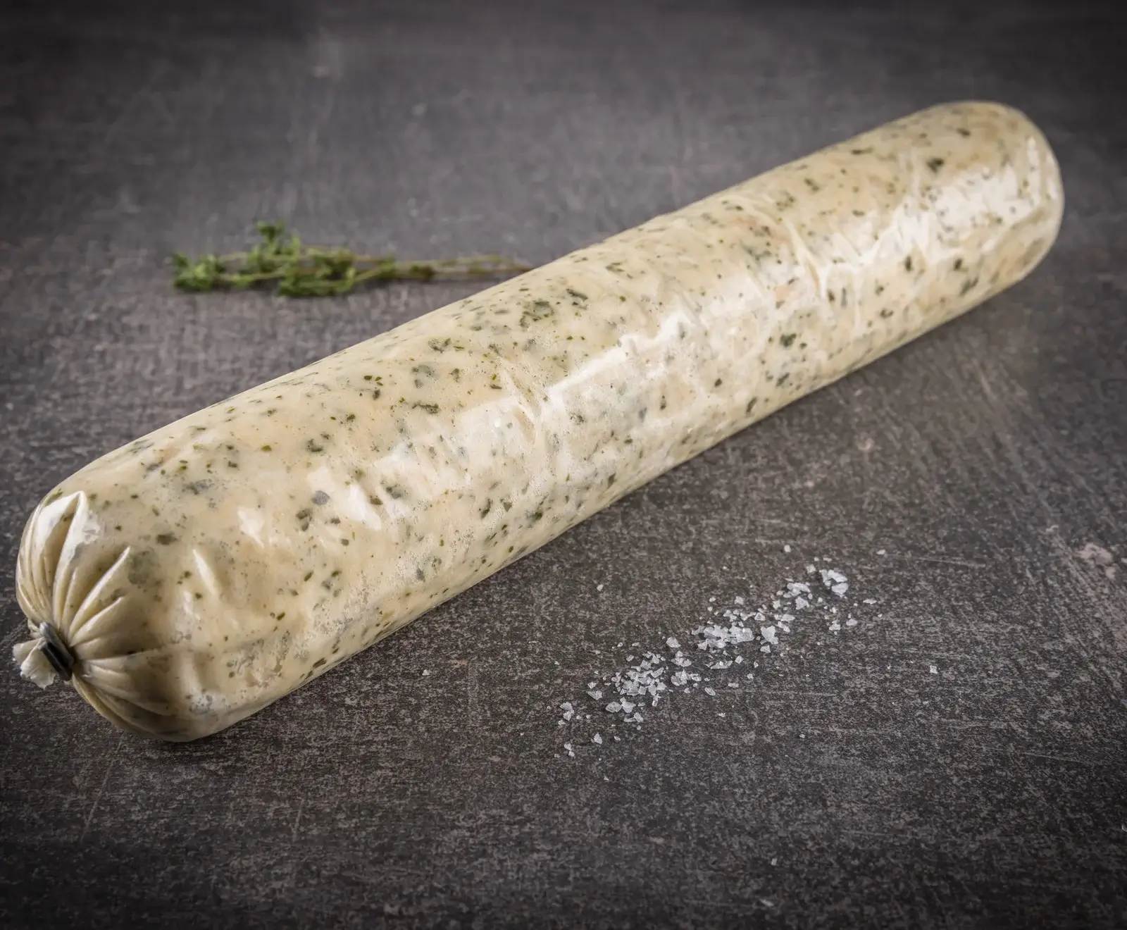 Eine in Klarsichtfolie verpackte Kräuterbutterrolle mit grünen Kräutersprenkeln liegt auf einem dunklen, strukturierten Untergrund. Ein Thymianzweig und grobes Salz sind ebenfalls sichtbar.