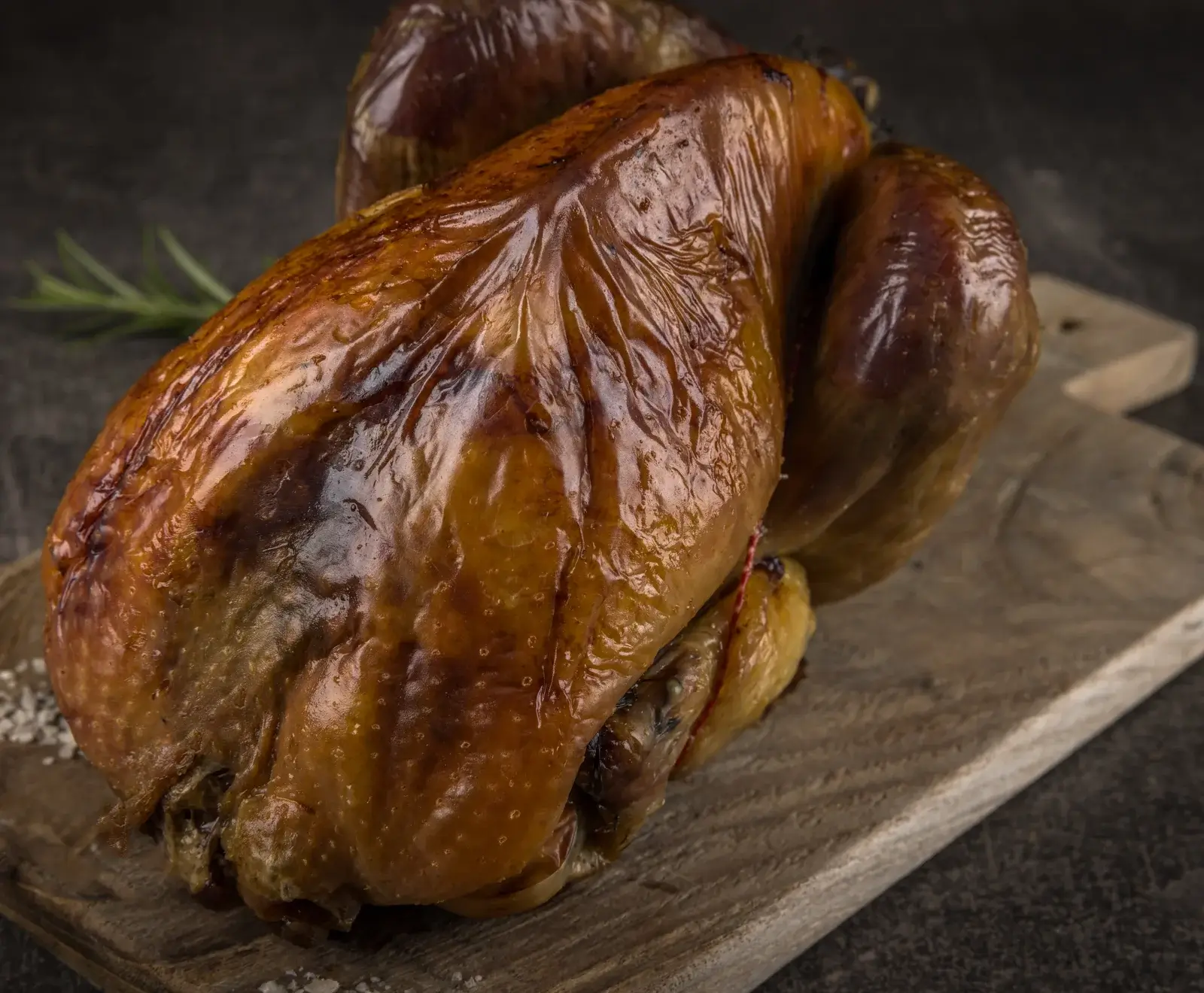 Ganzes, goldbraun glänzendes Brathähnchen auf einem rustikalen Holzbrett, garniert mit einem Rosmarinzweig und grobem Salz. Die Haut ist knusprig und appetitlich.