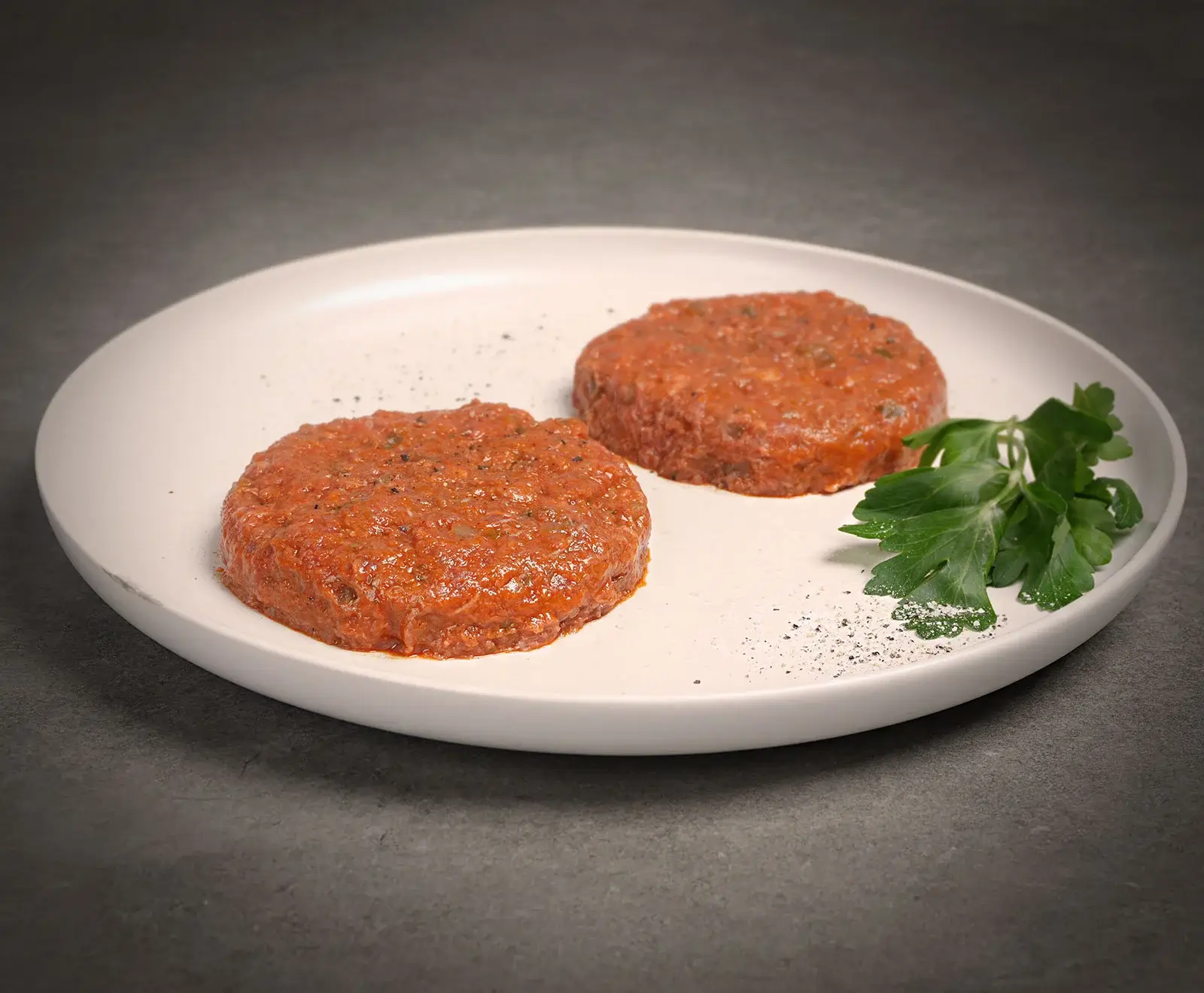 Zwei rohe, rötliche, gewürzte Patties auf einem weißen Teller, garniert mit frischer Petersilie und verstreuten Gewürzen. Der Hintergrund ist dunkelgrau.