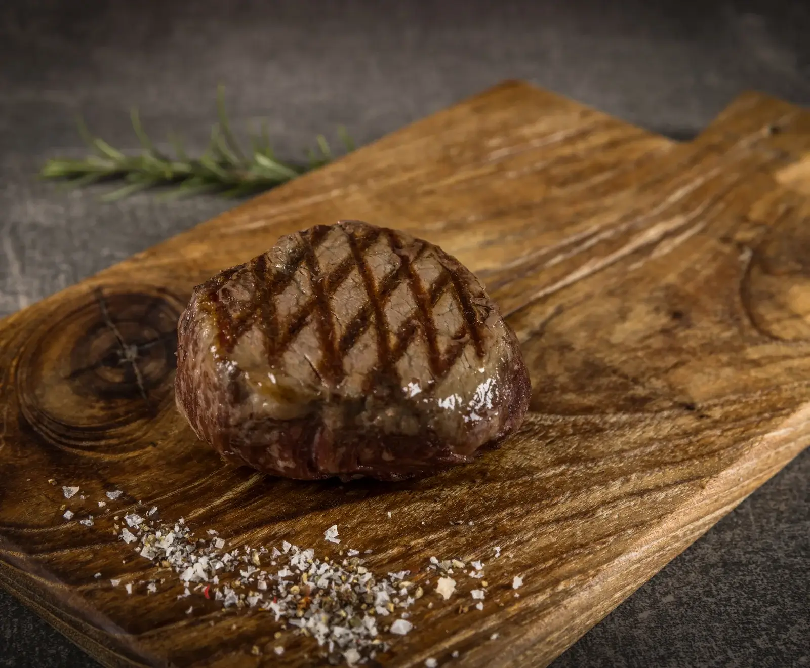 Ein perfekt gegrilltes Steak mit deutlichen Grillstreifen liegt auf einem rustikalen Holzschneidebrett. Davor sind grobes Salz und Pfeffer verstreut. Im Hintergrund ist ein Rosmarinzweig zu sehen.