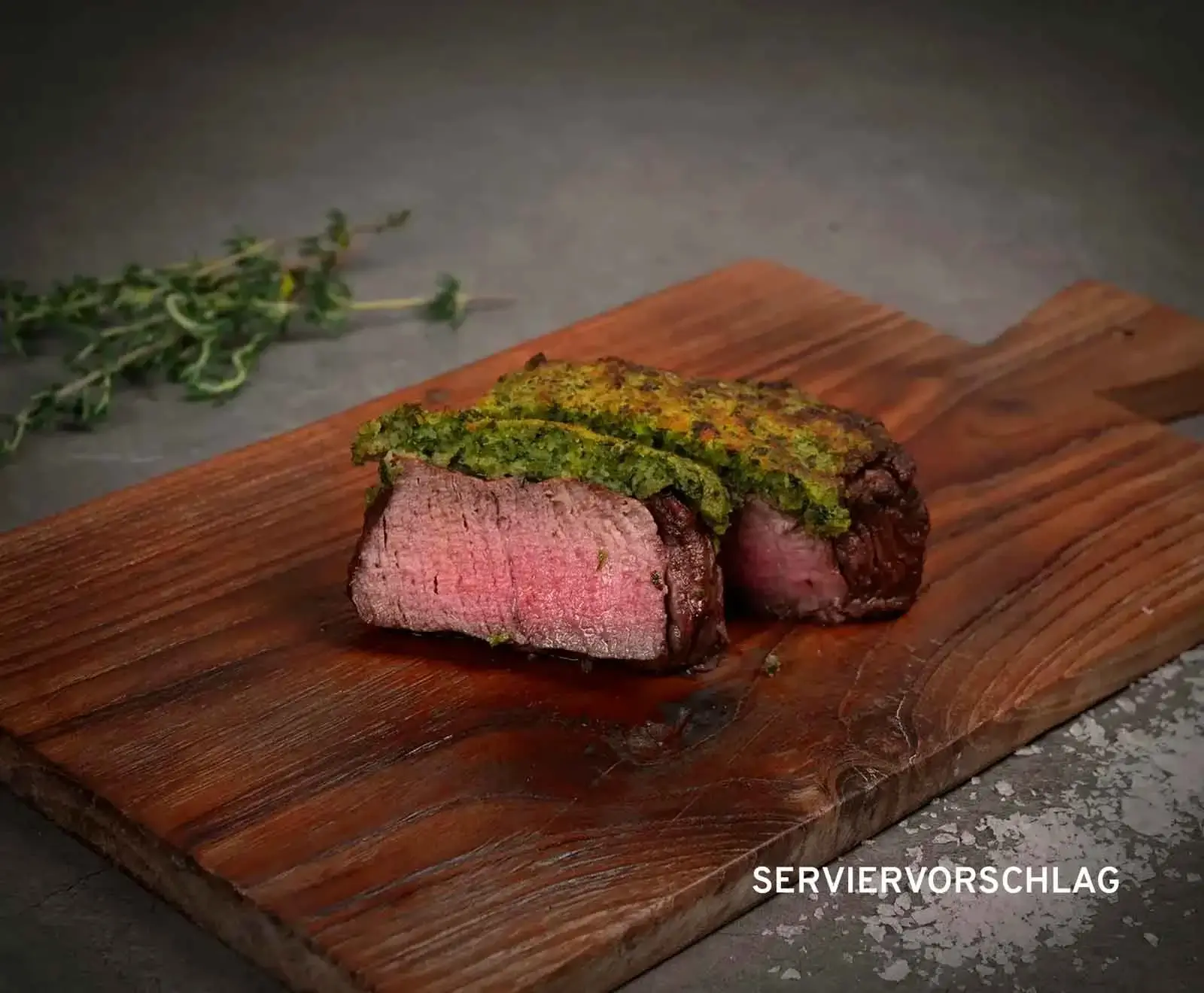 Zwei Stücke eines medium-rare gegarten Steaks mit grüner Kräuterkruste auf einem rustikalen Holzbrett. Im Hintergrund liegen frische Thymianzweige. Unten rechts steht "SERVIERVORSCHLAG".
