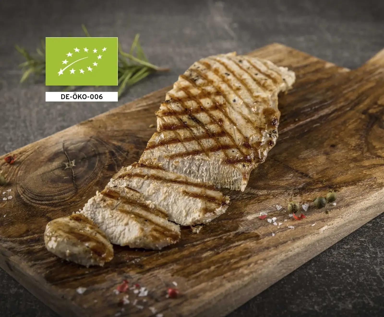 Gegrilltes, geschnittenes Bio-Hähnchenbrustfilet mit Grillstreifen auf einem Holzbrett, umgeben von Salz, Pfeffer und Rosmarin. Das EU-Bio-Logo ist sichtbar.