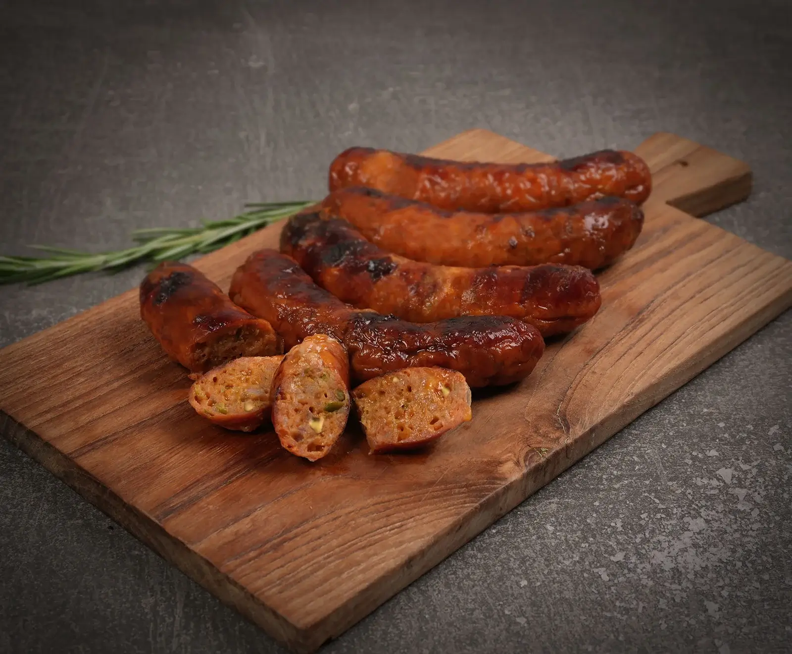 Mehrere gegrillte, braun gebratene Würste auf einem Holzschneidebrett. Eine Wurst ist in Scheiben geschnitten und zeigt die grobe Füllung mit grünen und hellen Stücken. Ein Rosmarinzweig liegt daneben.