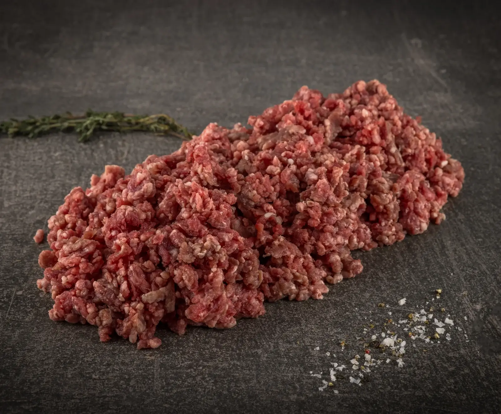 Ein großer Haufen frisches, rotes Hackfleisch liegt auf einer dunklen, strukturierten Oberfläche. Im Hintergrund ist ein Thymianzweig, im Vordergrund Salz- und Pfefferkörner.