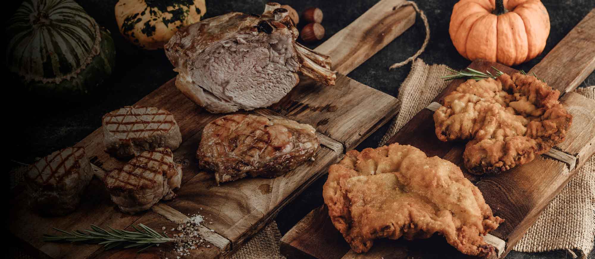 Verschiedene Fleischgerichte auf Holzbrettern: gegrillte Medaillons, ein großer Braten und knusprige panierte Schnitzel. Dekoriert mit Rosmarin, Salz, Pfeffer und herbstlichen Kürbissen auf dunklem Hintergrund.