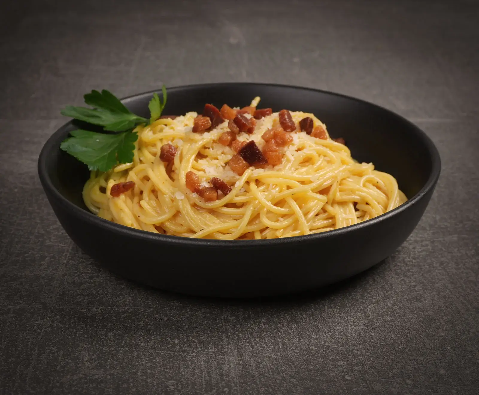 Eine schwarze Schale mit cremigen Spaghetti Carbonara, garniert mit knusprigen Speckwürfeln, geriebenem Käse und einem frischen Petersilienzweig, auf dunklem, strukturiertem Hintergrund.