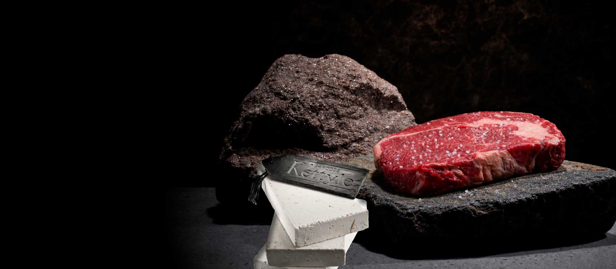 Ein rohes, gesalzenes Steak liegt auf einem dunklen Stein. Im Hintergrund ein großer, glitzernder Felsen, davor weiße Blöcke und ein schwarzes Etikett mit "Kettlye". Dunkle, rustikale Präsentation.