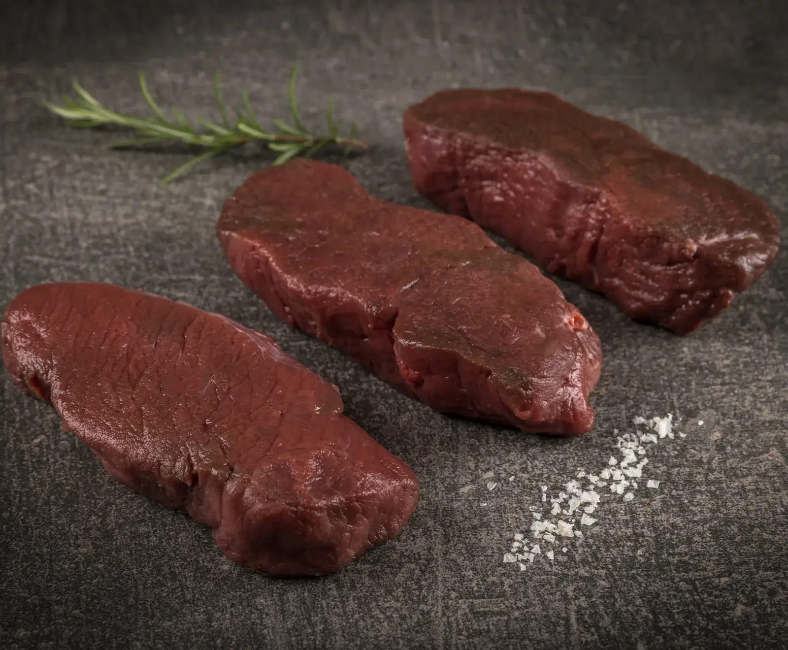 Drei rohe, dunkelrote Fleischstücke, vermutlich Wild oder Rind, liegen auf einer dunklen, strukturierten Steinplatte. Ein Rosmarinzweig und grobes Meersalz sind ebenfalls zu sehen.
