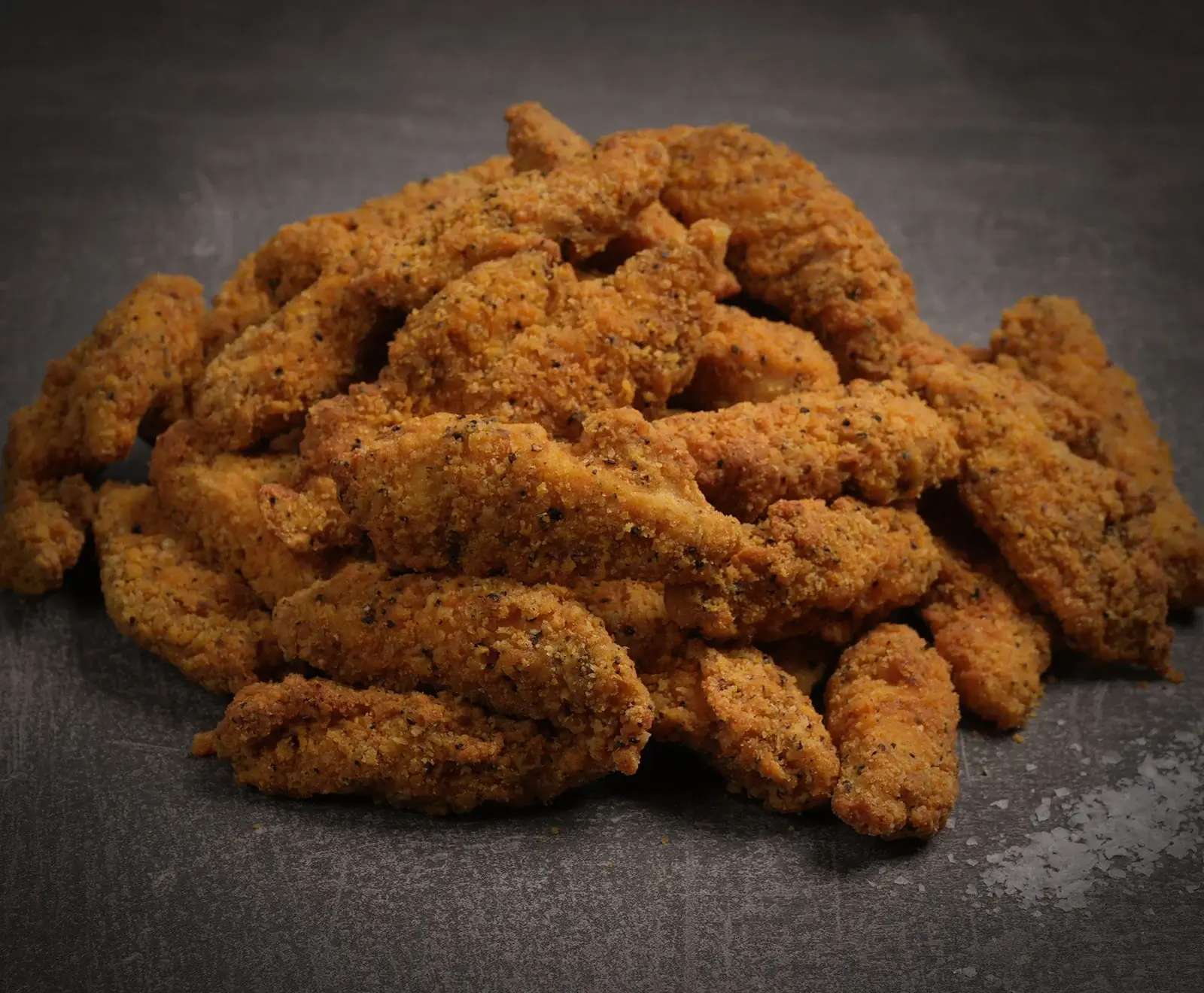 Ein Haufen goldbrauner, knusprig panierter Hähnchenstreifen mit dunklen Gewürzsprenkeln liegt auf einer dunklen, strukturierten Oberfläche.