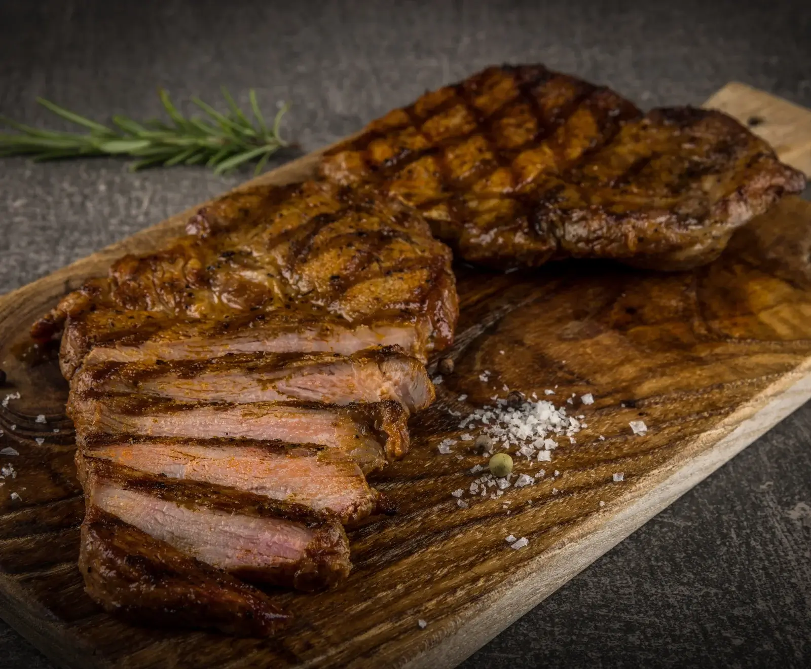 Saftige, gegrillte Steaks auf einem Holzbrett, eines geschnitten und medium-rare, mit Meersalz, Pfeffer und Rosmarin im Hintergrund.