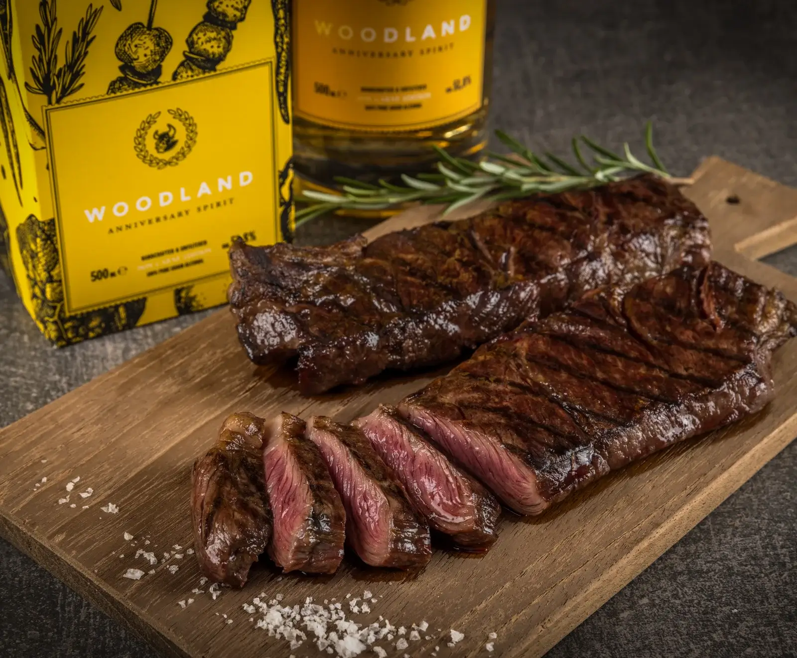 Zwei gegrillte Steaks auf einem Holzbrett, eines angeschnitten mit rosa Kern, daneben Meersalz. Im Hintergrund eine gelbe Flasche und Verpackung von "Woodland Anniversary Spirit" mit Rosmarin.