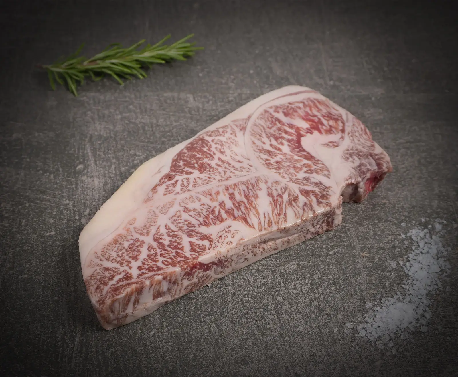 Ein rohes, stark marmoriertes Wagyu-Steak liegt auf einer dunklen Steinplatte, daneben ein frischer Rosmarinzweig und eine Prise grobes Meersalz.