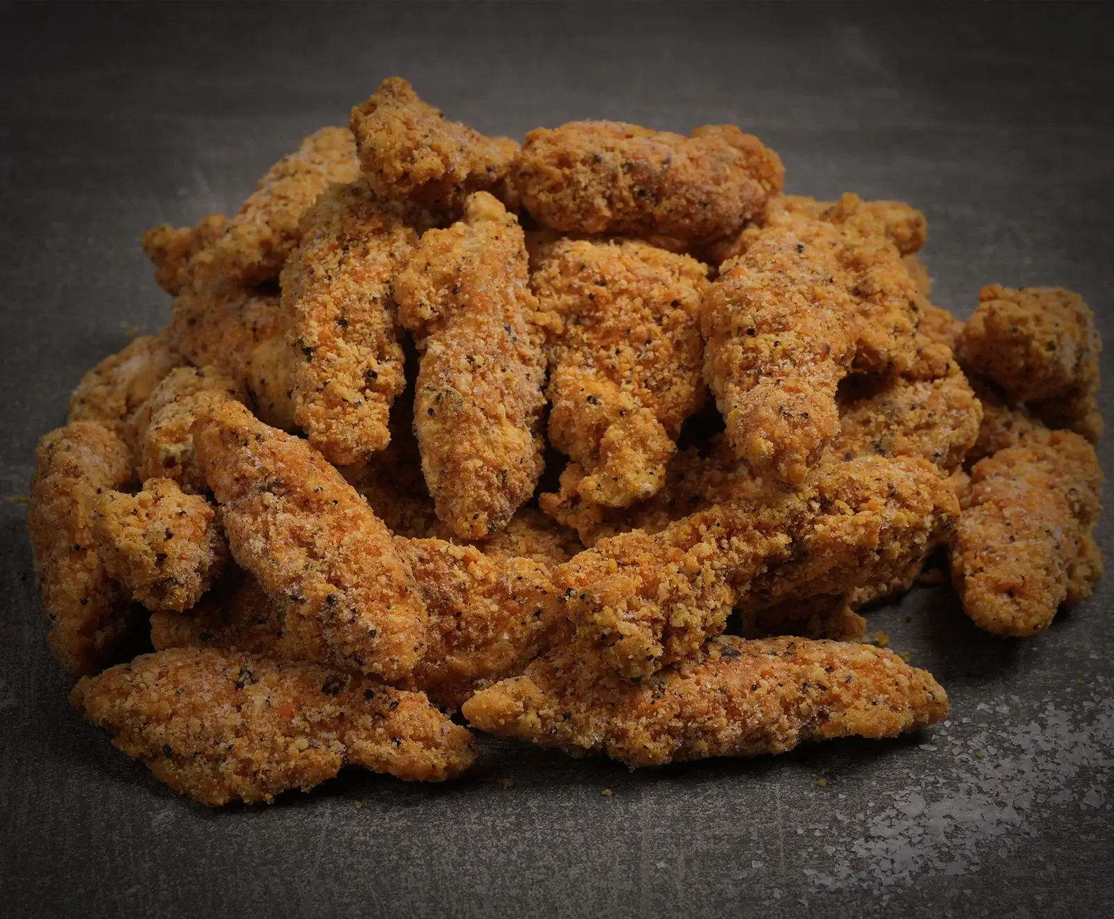 Ein Haufen ungekochter, panierter Hähnchen-Tender oder Nuggets mit einer groben, orange-braunen Panade und dunklen Gewürzsprenkeln liegt auf einem dunklen, strukturierten Untergrund.
