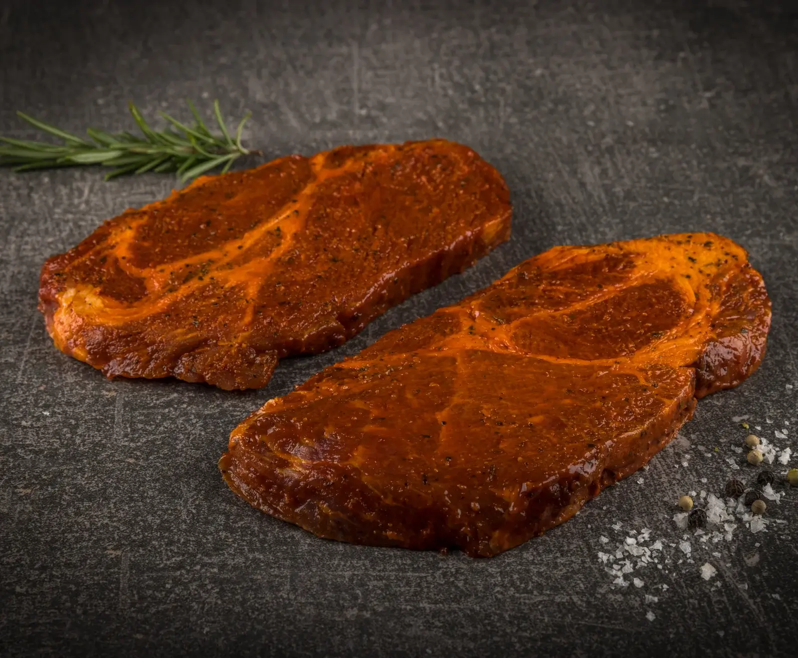 Zwei rohe, rot marinierte Schweinenackensteaks liegen auf einer dunklen Oberfläche, daneben ein Rosmarinzweig, grobes Salz und Pfefferkörner.