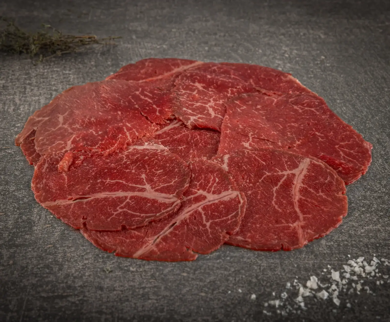 Dünne, rote, marmorierte Scheiben rohes Rindfleisch liegen übereinander auf einer dunklen Steinplatte. Im Hintergrund sind getrocknete Kräuter, im Vordergrund grobe Salzkristalle zu sehen.