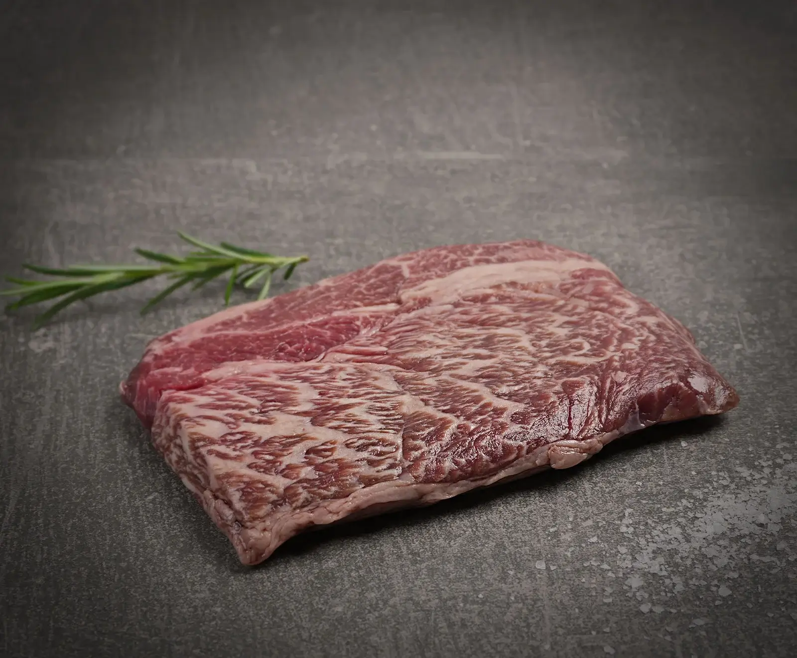 Rohes, stark marmoriertes Rindersteak auf dunklem, strukturiertem Untergrund, mit einem Rosmarinzweig im Hintergrund.