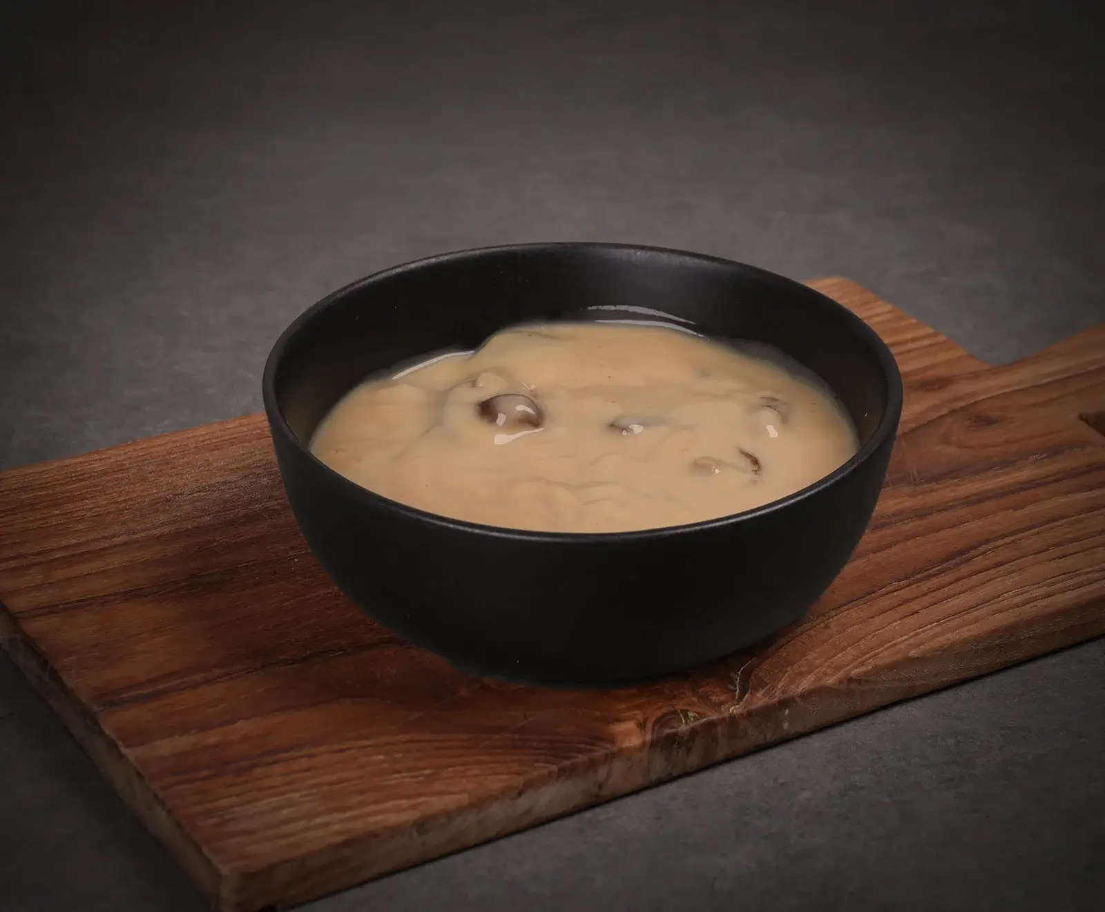 Eine schwarze Schale mit cremiger Pilzsauce, die kleine Pilzstücke enthält, steht auf einem rustikalen Holzbrett vor einem dunklen, grauen Hintergrund.