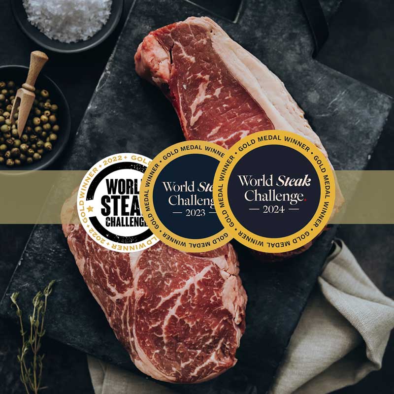 Zwei rohe Steaks auf dunklem Brett, umgeben von Salz, Pfefferkörnern und Rosmarin. Drei goldene "World Steak Challenge" Medaillen für 2022, 2023 und 2024 sind prominent platziert.