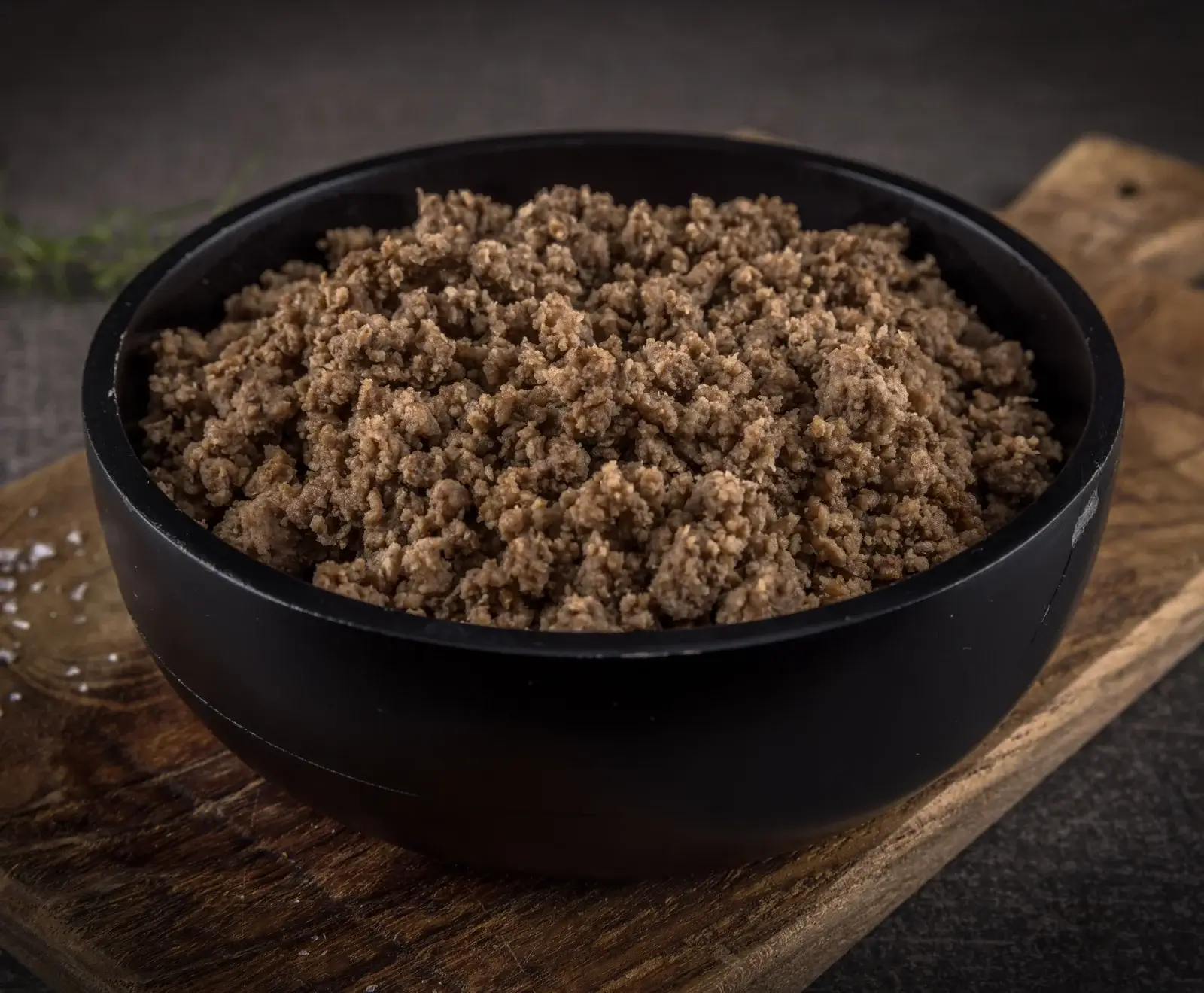 Eine schwarze Schüssel gefüllt mit braunem, gekochtem Hackfleisch, auf einem rustikalen Holzbrett. Im Hintergrund sind Kräuter und Salzkristalle zu sehen.