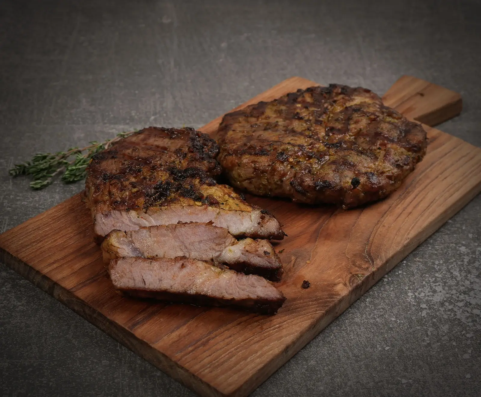 Zwei gegrillte Fleischstücke auf einem Holzschneidebrett. Ein Steak ist in Scheiben geschnitten und zeigt das rosa Innere, daneben liegt ein ganzes, rundes Fleischstück. Frischer Thymian ist im Hintergrund zu sehen.