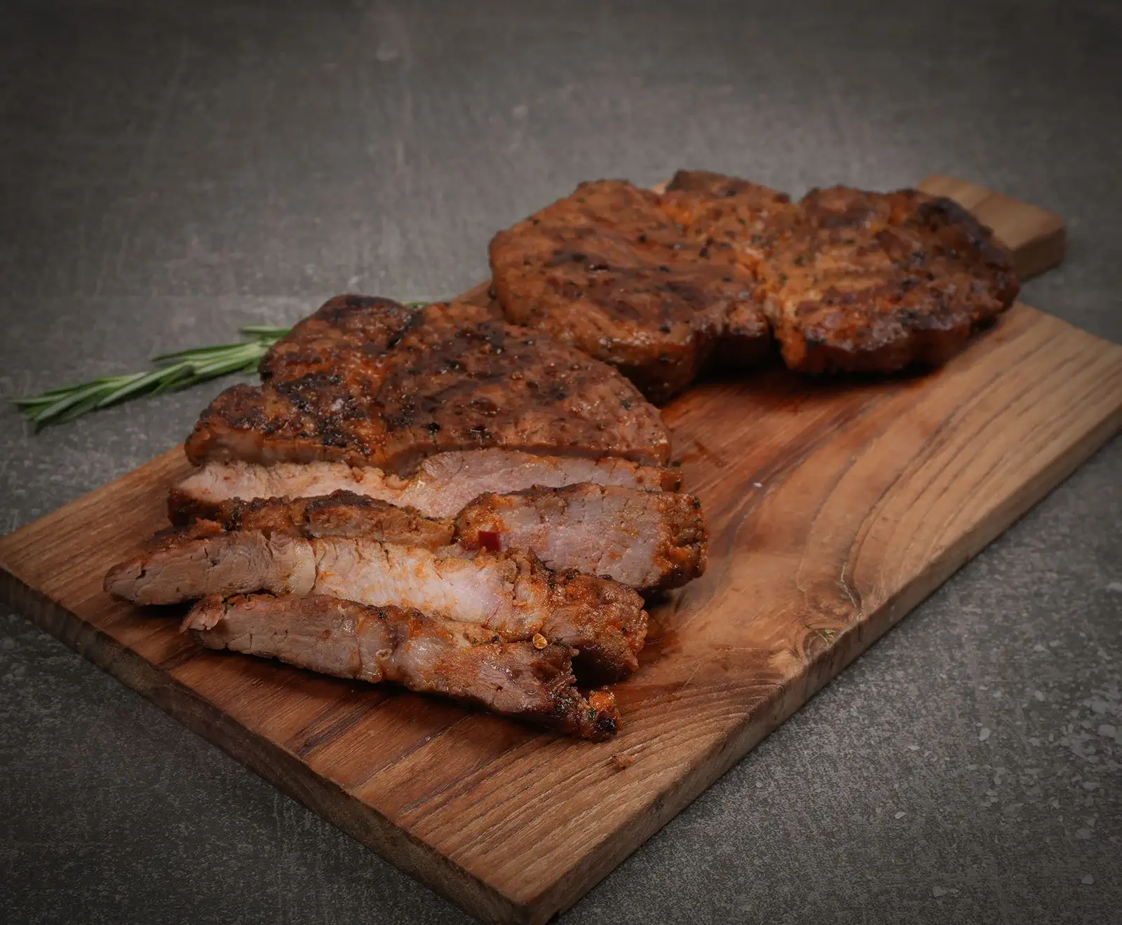 Zwei gegrillte Steaks auf einem Holzbrett, eines davon in Scheiben geschnitten, mit Rosmarin auf dunklem Hintergrund.
