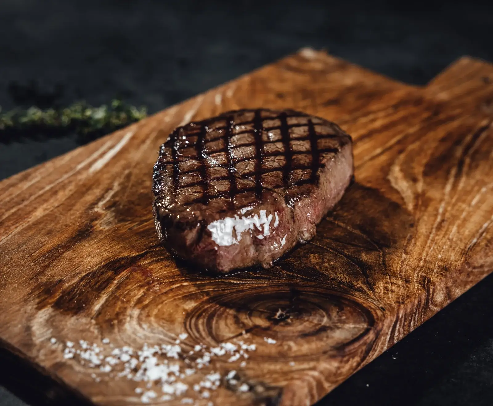 Ein perfekt gegrilltes Steak mit deutlichen Grillstreifen liegt auf einem rustikalen Holzschneidebrett. Grobes Meersalz ist verstreut, und im dunklen Hintergrund sind Kräuter zu sehen.
