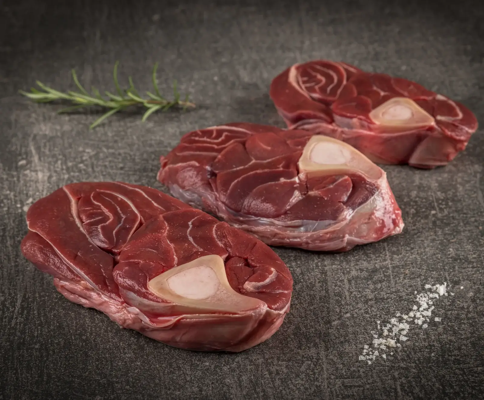 Drei rohe, dick geschnittene Fleischscheiben (vermutlich Ossobuco/Beinscheiben) mit Knochenmark in der Mitte, auf einer dunkelgrauen, strukturierten Oberfläche. Ein Rosmarinzweig und grobe Salzkristalle sind sichtbar.