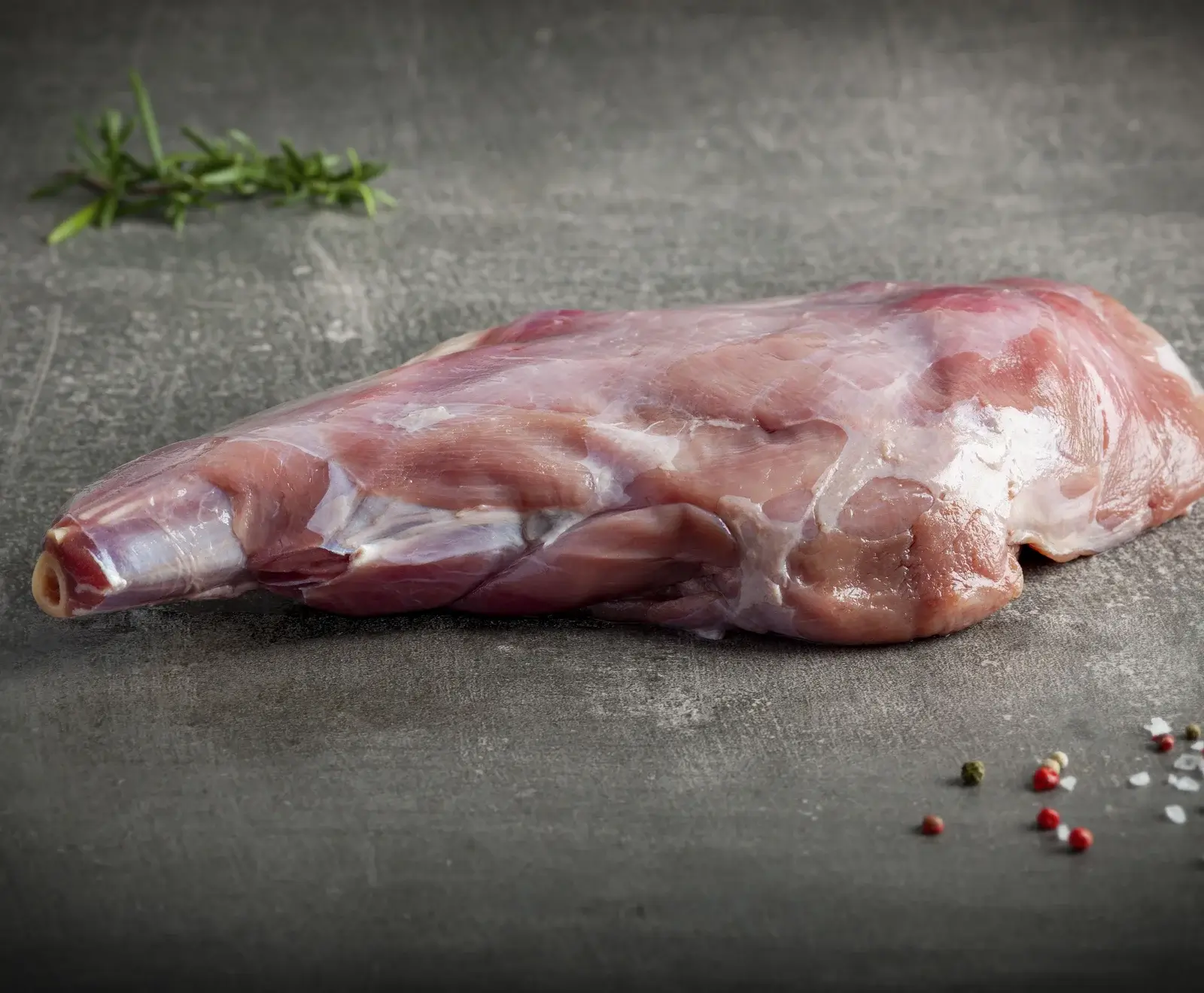 Ein rohes Stück Lamm- oder Kalbskeule liegt auf dunklem Untergrund. Im Hintergrund ist ein Rosmarinzweig, im Vordergrund sind Pfefferkörner und grobes Salz verstreut.