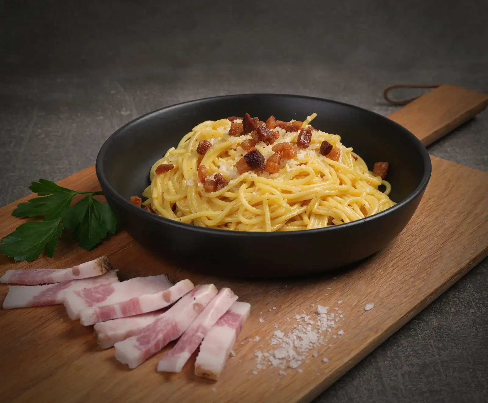 Spaghetti Carbonara in schwarzer Schale, garniert mit knusprigem Speck und geriebenem Käse. Auf einem Holzbrett liegen rohe Speckscheiben, Petersilie und grobes Salz.