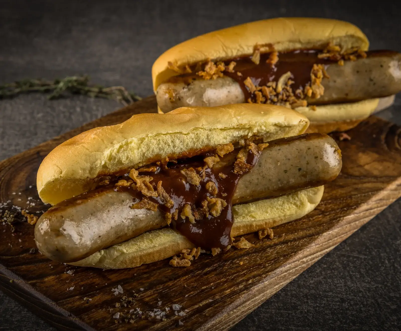 Zwei Bratwürste in Brötchen, übergossen mit dunkler Soße und Röstzwiebeln, auf einem Holzbrett mit grobem Salz und Rosmarin im dunklen Hintergrund.