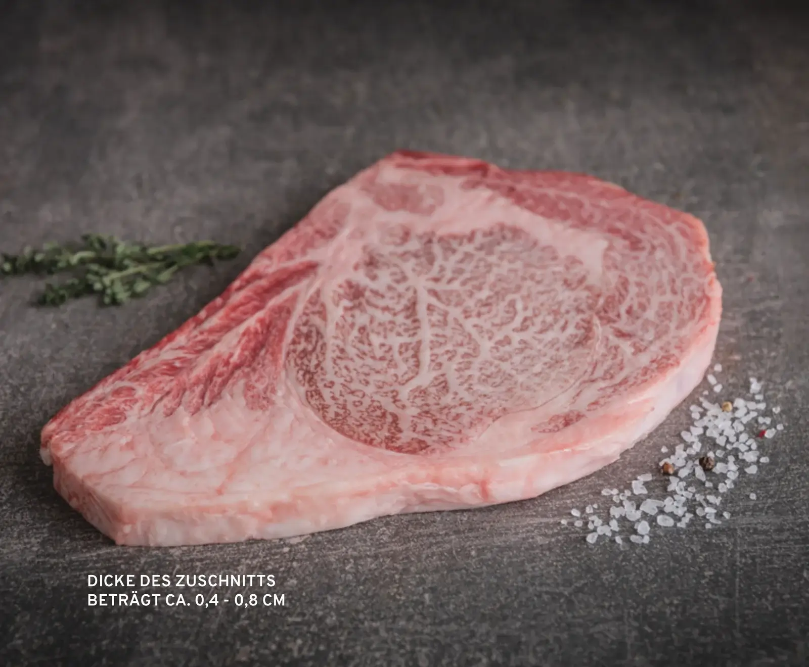 Ein rohes, stark marmoriertes Stück Rindfleisch, vermutlich Wagyu, liegt auf einer dunklen Oberfläche. Daneben sind grobes Salz, Pfeffer und ein Thymianzweig zu sehen. Die Schnittdicke beträgt ca. 0,4 - 0,8 cm.