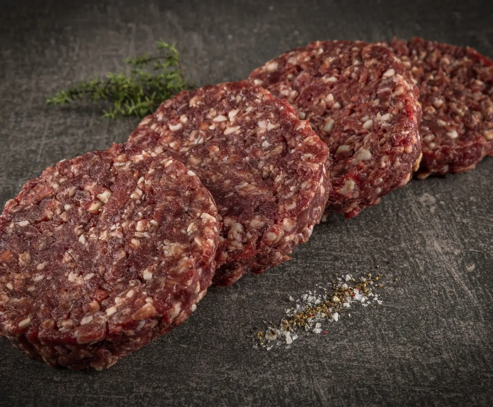 Vier rohe, runde Burger-Patties aus Hackfleisch liegen diagonal auf einer dunklen Oberfläche. Im Hintergrund ist ein Thymianzweig, im Vordergrund Salz und Pfeffer.