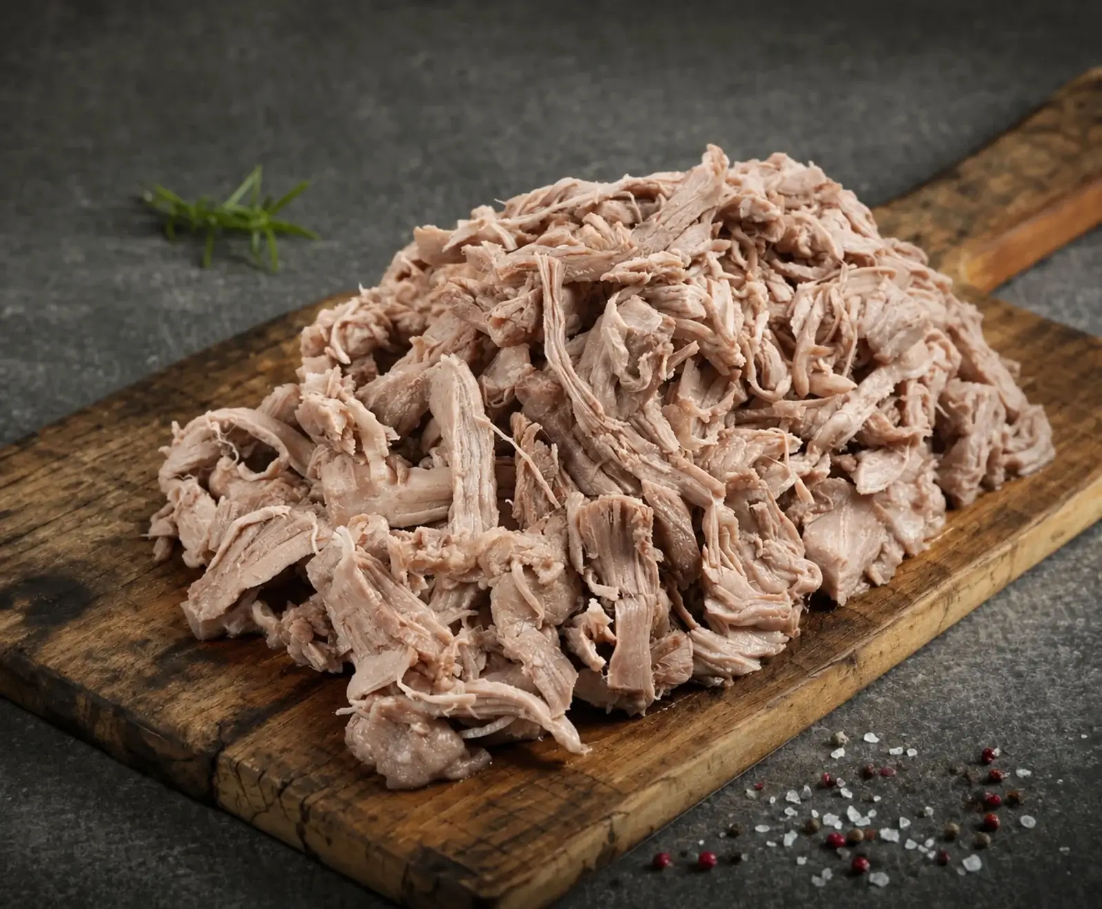 Ein großer Haufen zerkleinertes, hellbraunes Fleisch (Pulled Pork) auf einem rustikalen Holzbrett. Im Hintergrund ein Rosmarinzweig, im Vordergrund verstreute Salz- und Pfefferkörner auf dunklem Untergrund.
