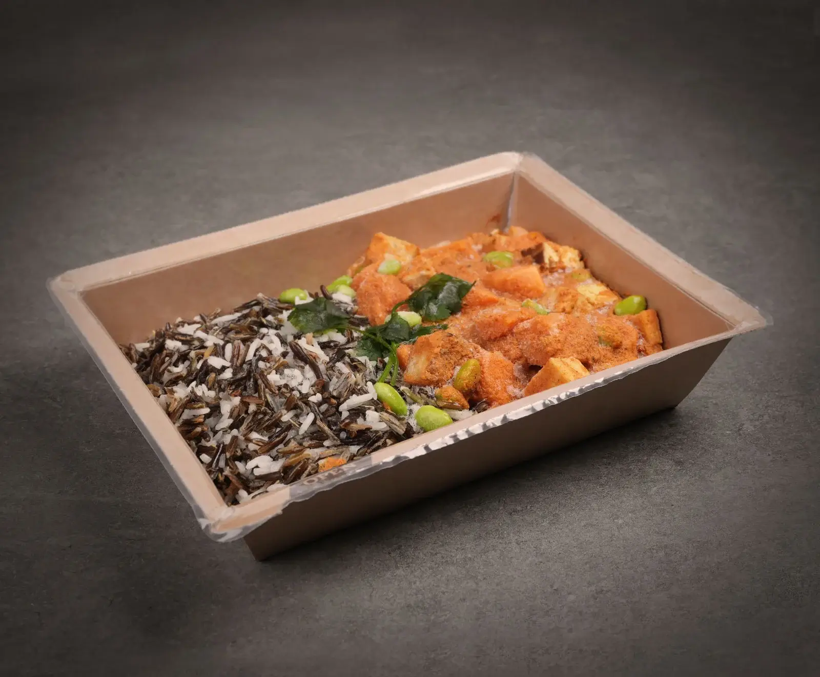 Eine rechteckige, braune Pappschale mit durchsichtigem Deckel, gefüllt mit einer Mahlzeit. Links eine Mischung aus Wildreis und weißem Reis, rechts gewürfeltes Essen in orangeroter Soße, garniert mit Edamame und grünen Kräutern auf dunklem Untergrun