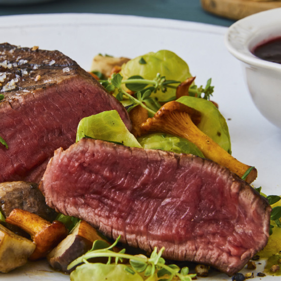 Nahaufnahme eines aufgeschnittenen, medium-rare Steaks, mit grobem Salz bestreut. Serviert mit Pfifferlingen, weiteren Pilzen, grünen Blättern und frischen Kräutern auf einem weißen Teller. Eine Schale mit dunkler Soße ist im Hintergrund.