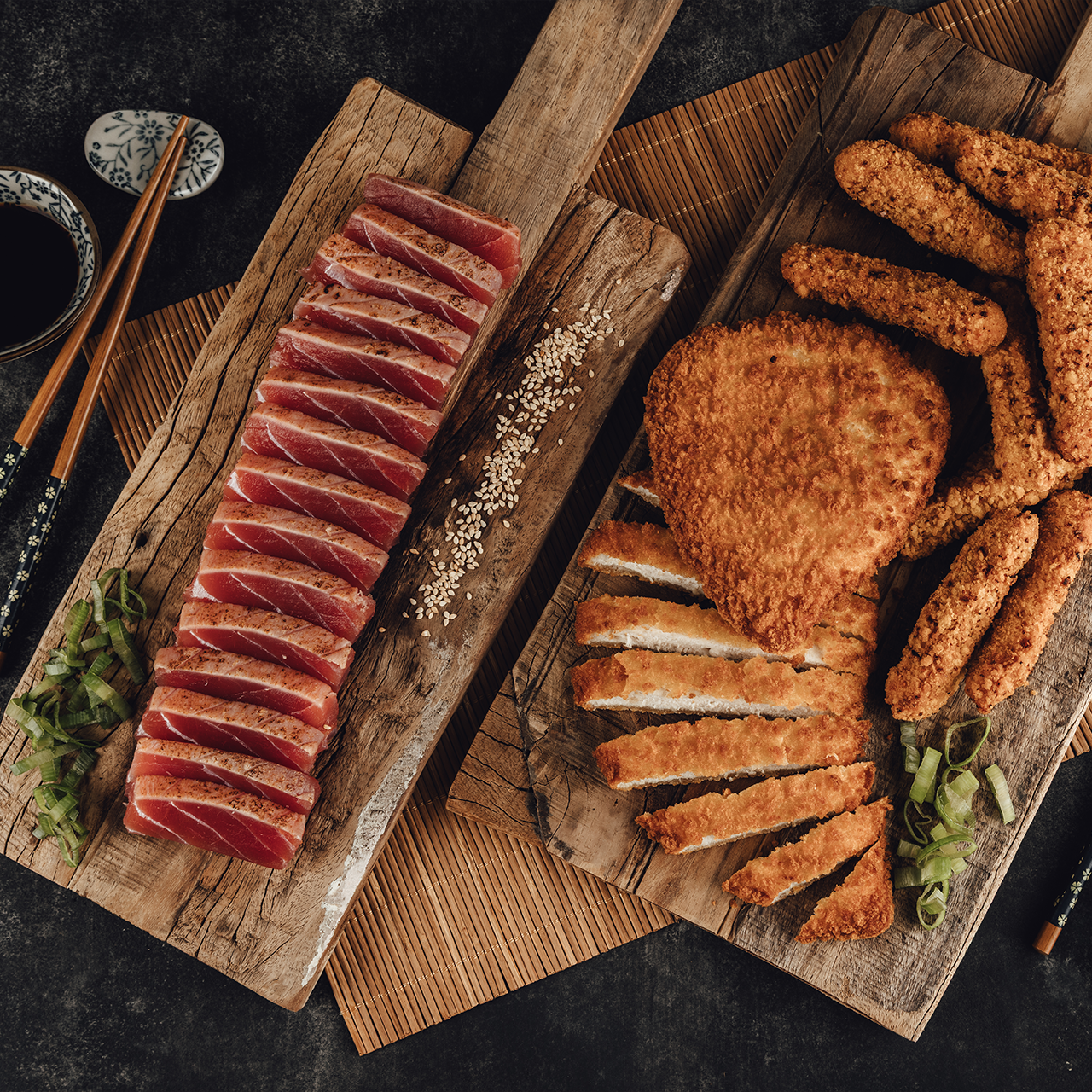 Draufsicht auf zwei Holzbretter mit japanischen Gerichten: Links geschnittener, leicht angebratener Thunfisch mit Sesam und Frühlingszwiebeln, daneben Sojasauce und Essstäbchen. Rechts paniertes Schnitzel und frittierte Streifen mit Frühlingszwiebeln.