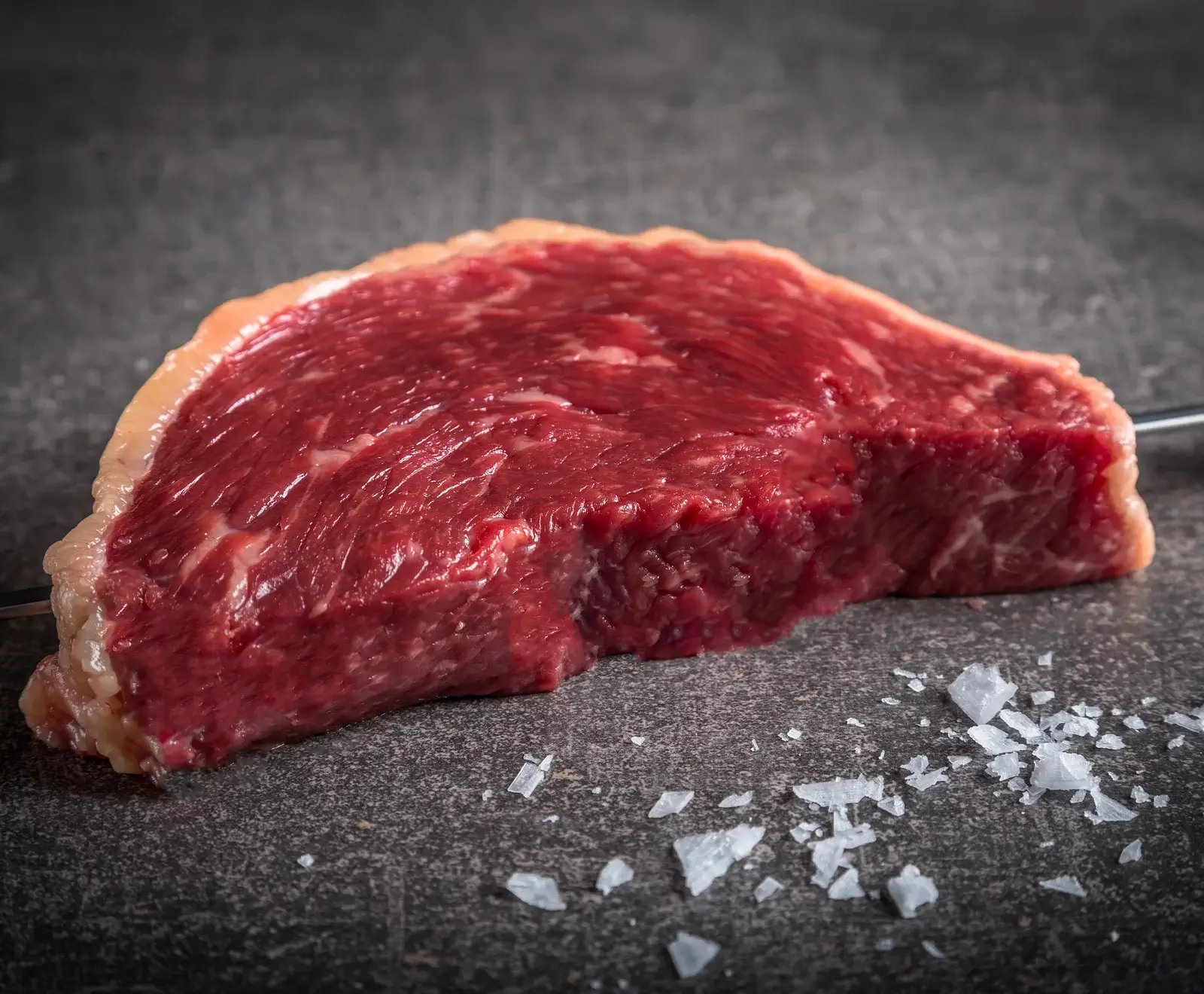 Rohes Picanha-Steak mit einer dicken Fettschicht liegt auf dunklem Untergrund, daneben grobe Meersalzflocken. Ein Metallspieß ist durch das Fleisch gesteckt.