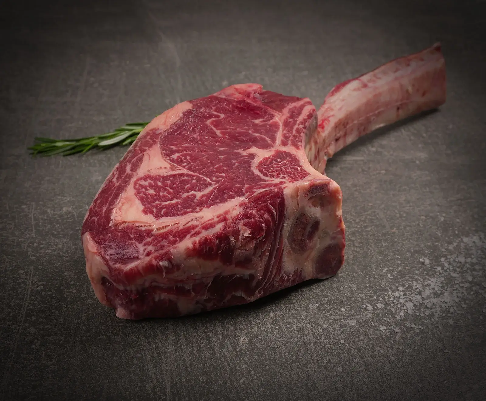 Ein rohes Tomahawk-Steak mit langem Knochen und schöner Marmorierung liegt auf einer dunklen, strukturierten Oberfläche. Ein Rosmarinzweig ist im Hintergrund zu sehen.