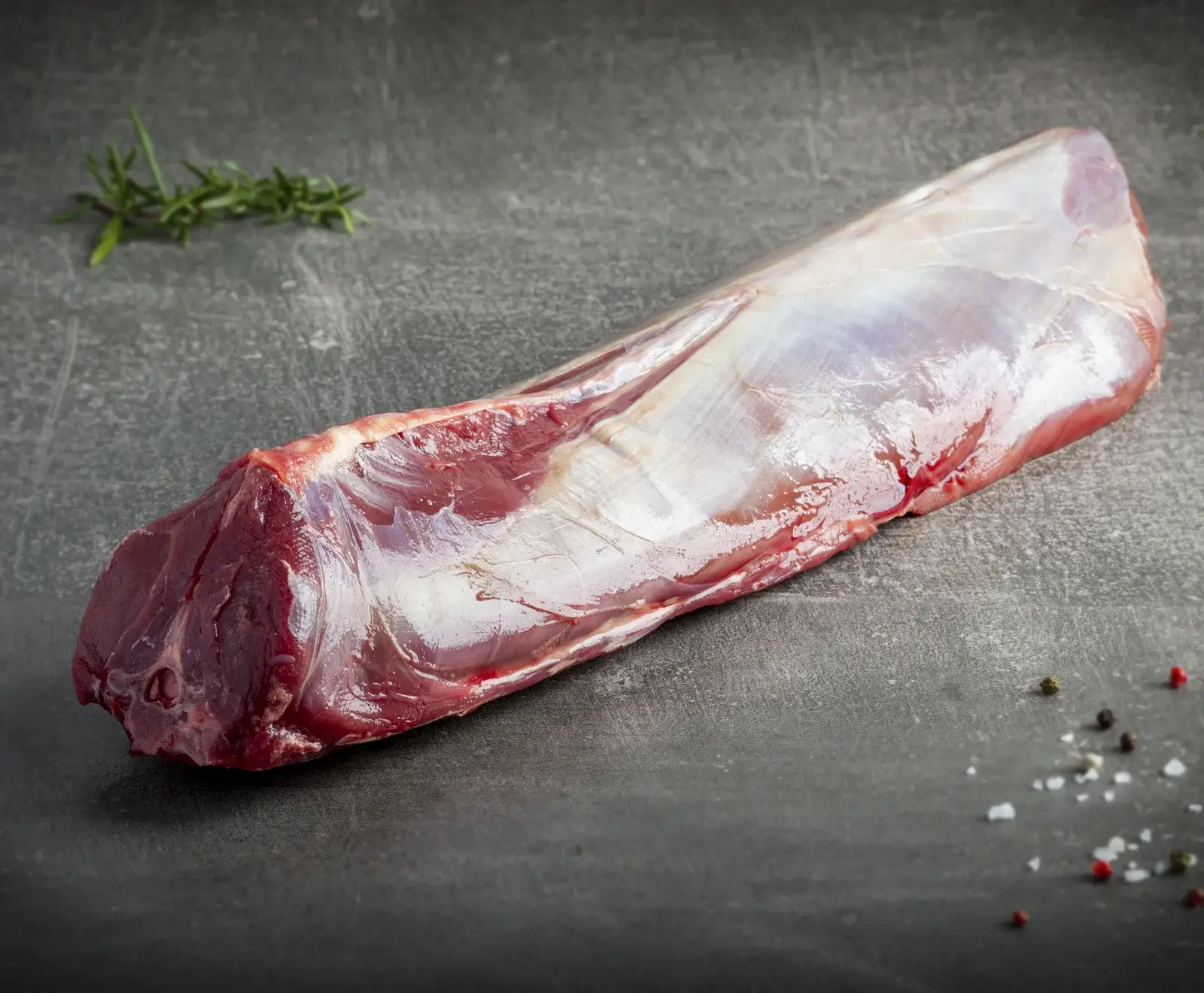 Ein frisches, rohes Stück Fleisch, vermutlich Wild- oder Rinderfilet, mit Silberhaut auf dunklem Untergrund. Daneben liegen Rosmarinzweige, bunte Pfefferkörner und grobes Meersalz.