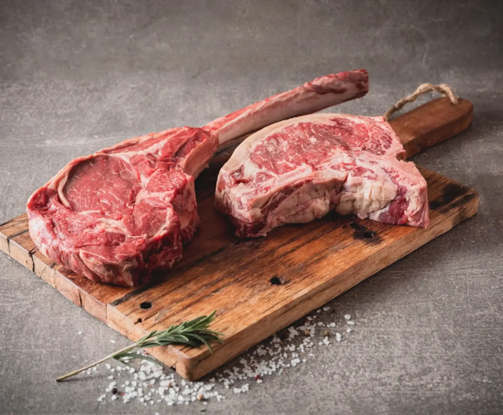 Zwei rohe Rindersteaks auf einem hölzernen Schneidebrett mit Salz und einem Rosmarinzweig.