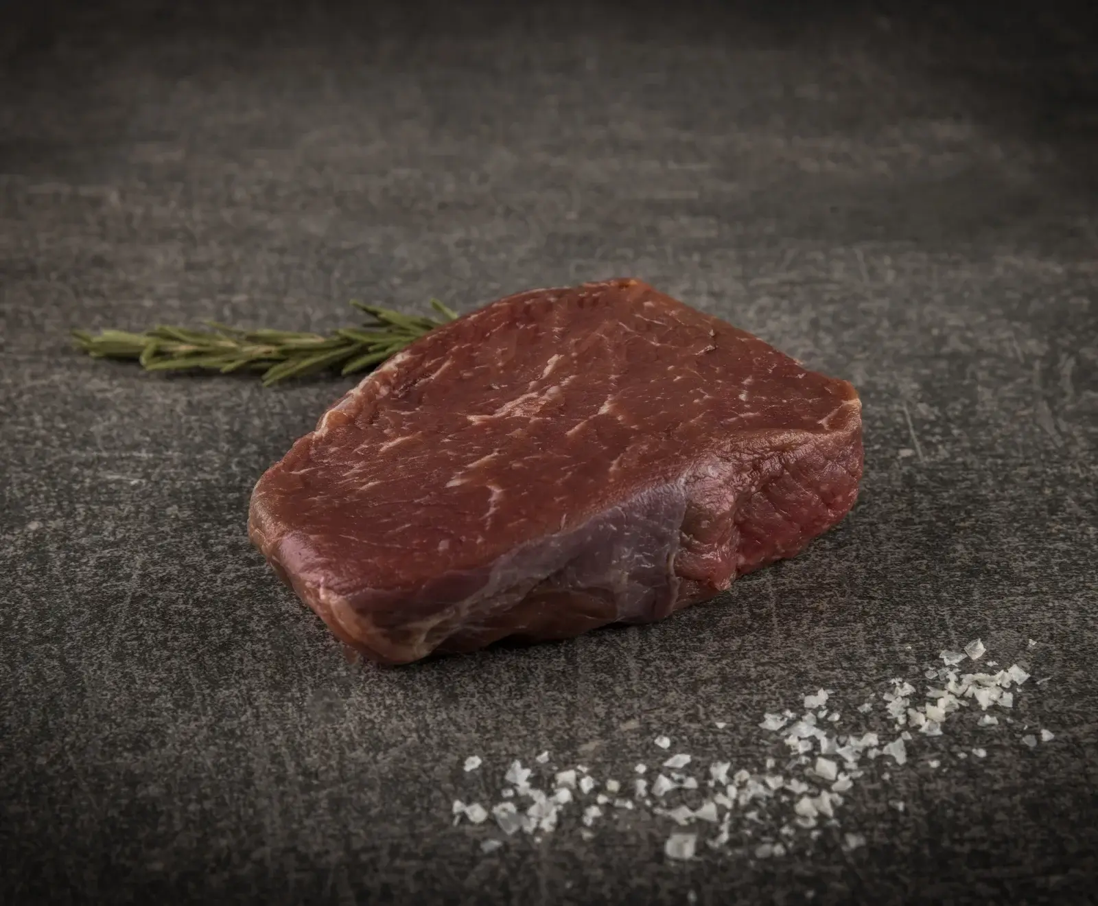 Ein rohes, rotes Steak liegt auf einer dunklen Steinplatte. Dahinter befindet sich ein Rosmarinzweig, davor grobes Meersalz. Das Fleisch ist leicht marmoriert.