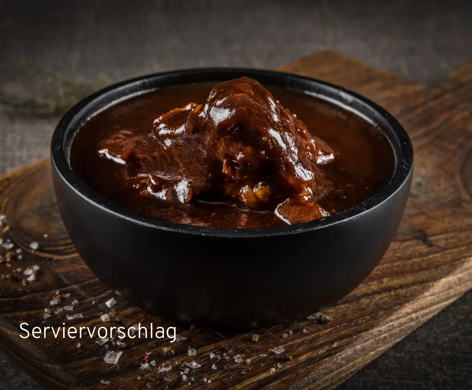 Eine schwarze Schale mit glänzendem, dunklem Gulasch oder Schmorgericht, serviert auf einem rustikalen Holzbrett mit grobem Salz. Im Vordergrund steht der Text "Serviervorschlag".