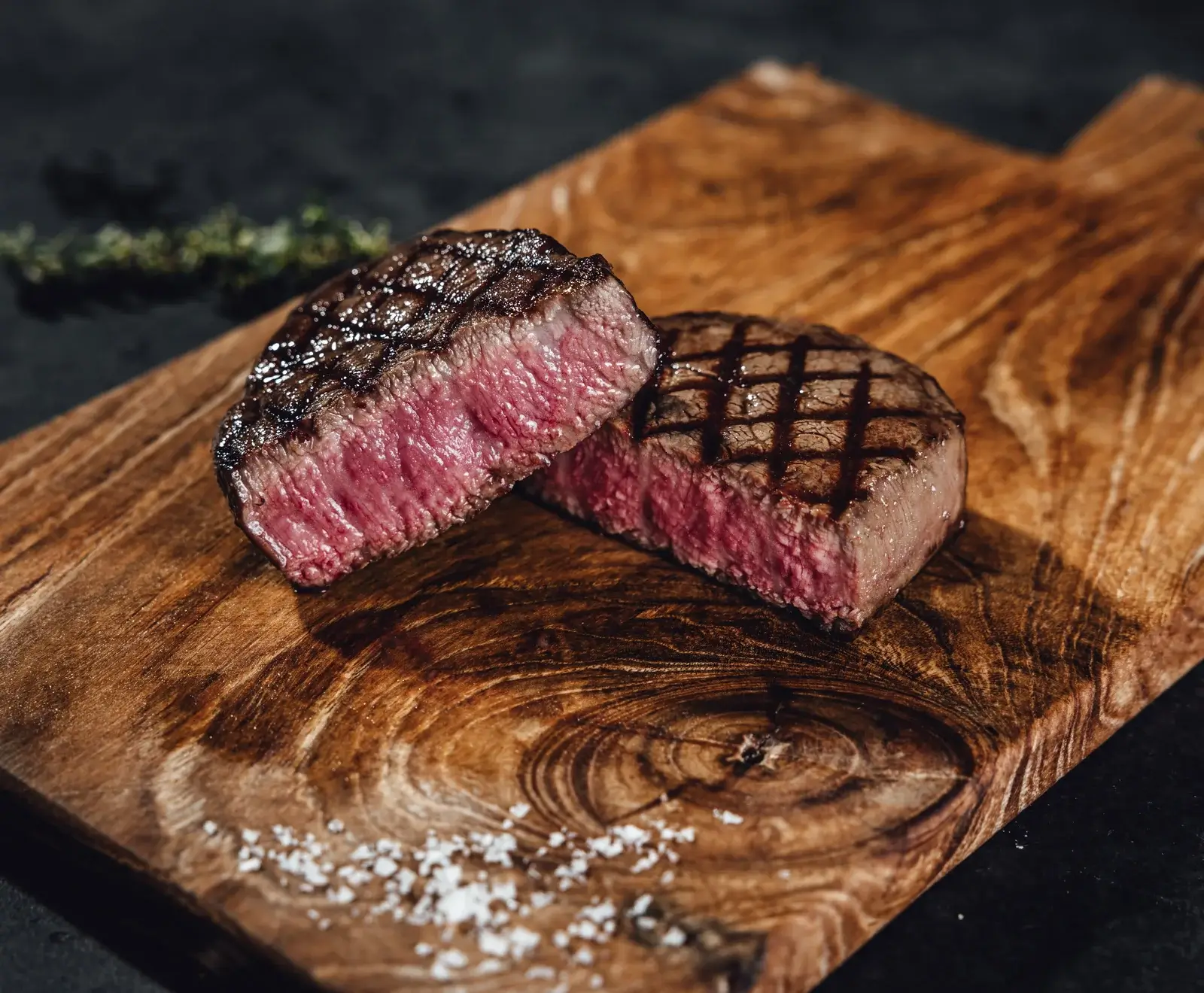 Zwei gegrillte Steaks auf einem rustikalen Holzbrett, eines halbiert mit rosa Kern und deutlichen Grillstreifen. Grobes Meersalz und ein Kräuterzweig sind ebenfalls sichtbar.